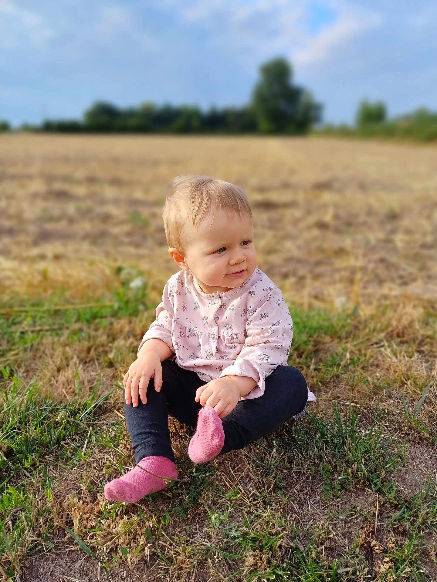 Thalia a rejoint le concours — aidez-le/la à gagner de superbes lots ! toddler, child, grass, field, outdoor, nature, pink_socks, pink_top, curious, sitting, baby, portrait, daylight, casual_clothing, nature_background, young_child, blond_hair, looking_away, soft_light, rural
