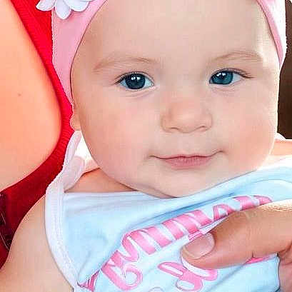 Athena joined the competition — help win amazing prizes! baby, infant, child, portrait, close_up, face, smiling, blue_eyes, floral_headband, flowers, pink, pastel, bib, sleeveless_outfit, adult_hands, holding, indoor, photograph, cute, family