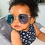 Lila participe au concours pour gagner de l'argent avec cette photo : baby, child, curly_hair, sunglasses, high_chair, floral_outfit, indoors, face, portrait, cute, skin, arm, table, expression, person, fashion, eyewear, seat, infant, closeup