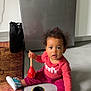 Lila a rejoint le concours — aidez-le/la à gagner de superbes lots ! toddler, child, indoor, floor, toy, stacking_cups, red_shirt, blue_shoes, curly_hair, kitchen, refrigerator, wooden_spoon, colorful, rug, bag, person, sitting, cute, playful, expression