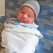 Charlie is registered to the contest to win money with this photo: newborn, baby, infant, sleeping, swaddled, blanket, hat, gray_hat, white_clothing, portrait, face, closed_eyes, peaceful, cozy, indoors, textile, patterned_background, sitting, resting, photography