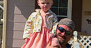 Dalilah is registered to the contest to win money with this photo: child, man, outdoor, house, porch, fence, sunglasses, cap, dress, floral_jacket, smile, grass, daylight, holding, person, window, shirt, beard, footwear, shadow
