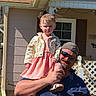 child, man, outdoor, house, porch, fence, sunglasses, cap, dress, floral_jacket, smile, grass, daylight, holding, person, window, shirt, beard, footwear, shadow