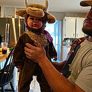 Oliver is registered to the contest to win money with this photo: adult, baby, beverage, bull_costume, casual, chair, child, costume, cute, furniture, holding, home, indoor, kitchen, person, plush, portrait, refrigerator, smile, table