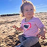 toddler, child, beach, sand, sunlight, curly_hair, pink_shirt, nike_logo, leggings, barefoot, outdoor, daytime, playful, portrait, sitting, casual_clothing, sunny, young_child, nature, curious_expression