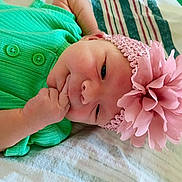 Rylee is registered to the contest to win money with this photo: baby, infant, child, portrait, lying_down, bed, green_outfit, pink_flower, headband, floral_accessory, rosy_cheeks, smiling, hand, fist, indoor, close_up, colorful, soft_lighting, blanket, photography