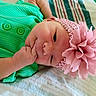 baby, infant, child, portrait, lying_down, bed, green_outfit, pink_flower, headband, floral_accessory, rosy_cheeks, smiling, hand, fist, indoor, close_up, colorful, soft_lighting, blanket, photography