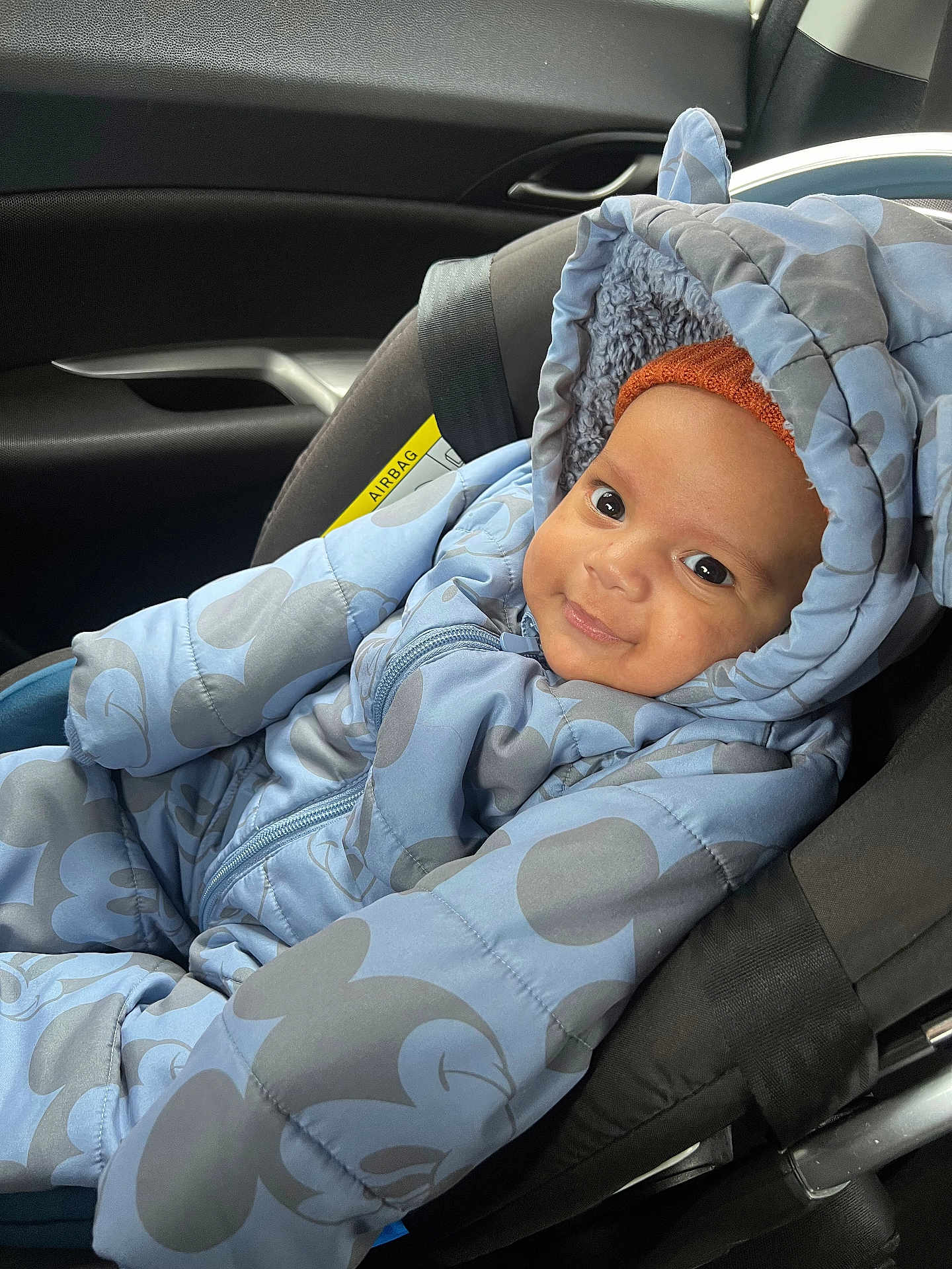 Leon is registered to the contest to win money with this photo: baby, child, car_seat, snowsuit, blue_clothing, mickey_mouse_pattern, seatbelt, smile, face, headwear, orange_hat, indoors, car_interior, window, safety, cute, warm_clothing, infant, portrait, child_safety
