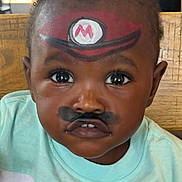 Kentrell joined the competition — help win amazing prizes! child, toddler, face_paint, mario_hat, mustache_paint, curly_hair, big_eyes, shirt, cartoon_character, indoor, portrait, closeup, playful, cute, person, expression, colorful, young_child, casual_clothing, wooden_background