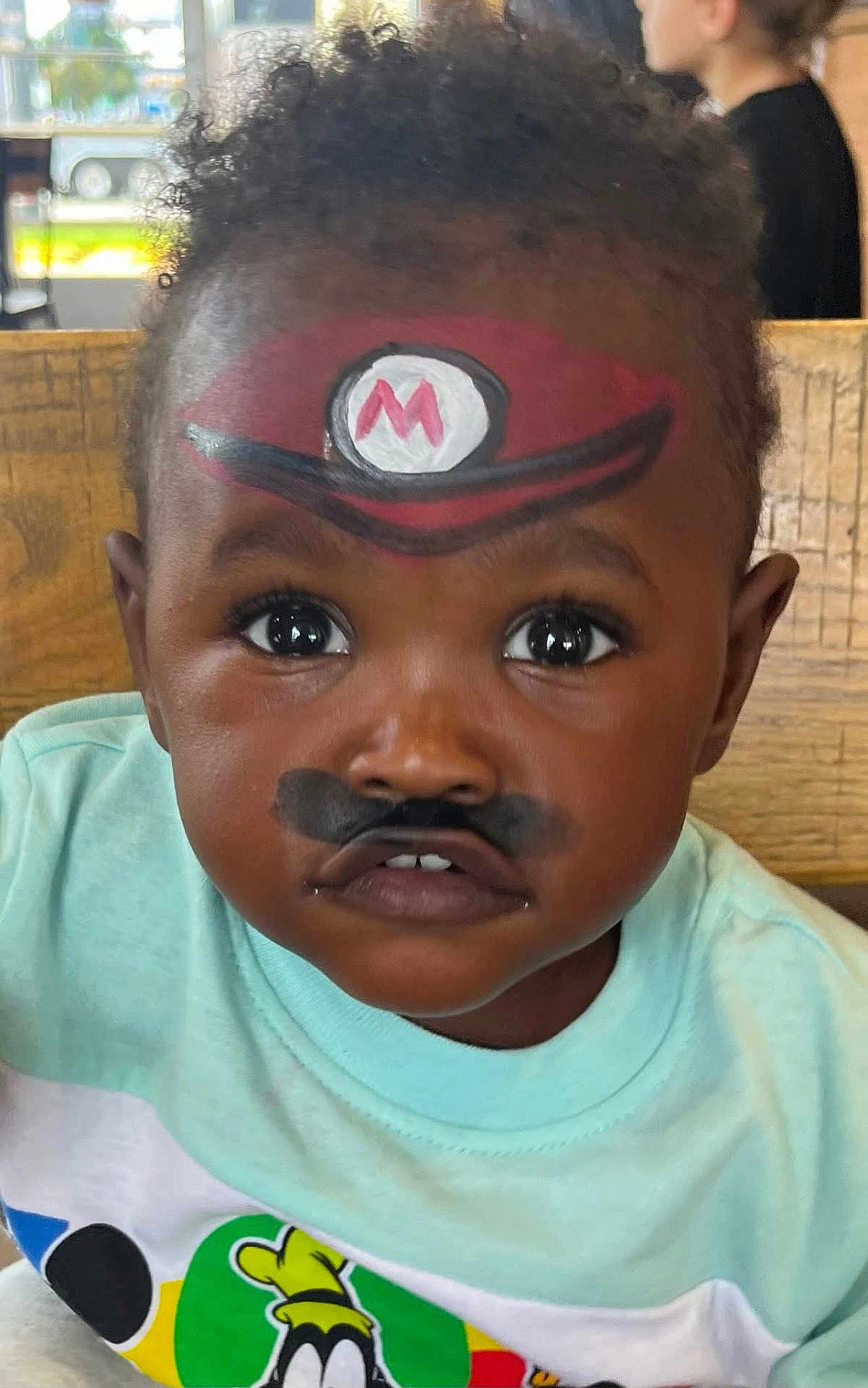 Kentrell joined the competition — help win amazing prizes! child, toddler, face_paint, mario_hat, mustache_paint, curly_hair, big_eyes, shirt, cartoon_character, indoor, portrait, closeup, playful, cute, person, expression, colorful, young_child, casual_clothing, wooden_background