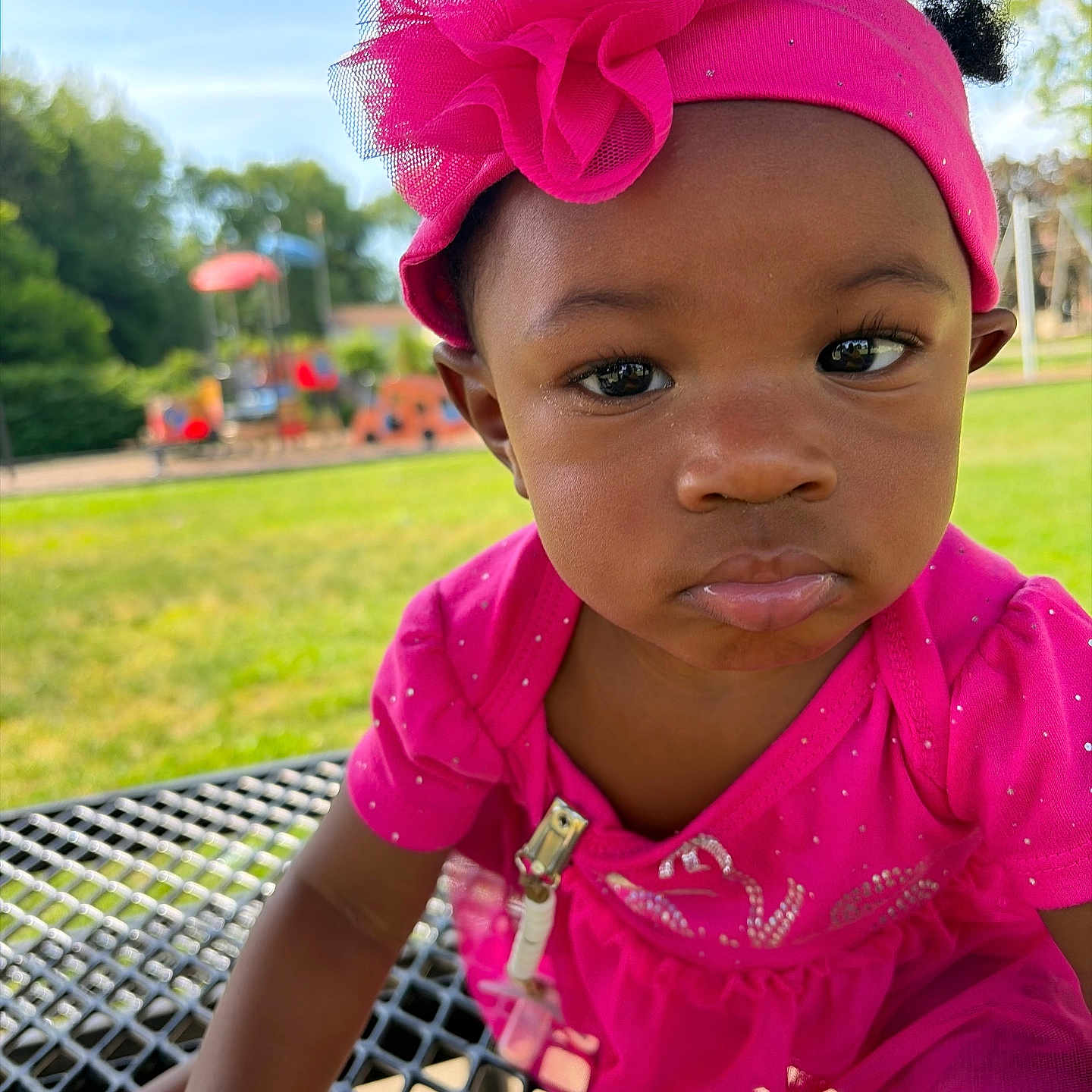 Iomi joined the competition — help win amazing prizes! toddler, child, pink_dress, headband, flower, outdoor, playground, grass, table, metal_surface, face, expression, curious, sunlight, daylight, person, young_child, cute, close_up, summer