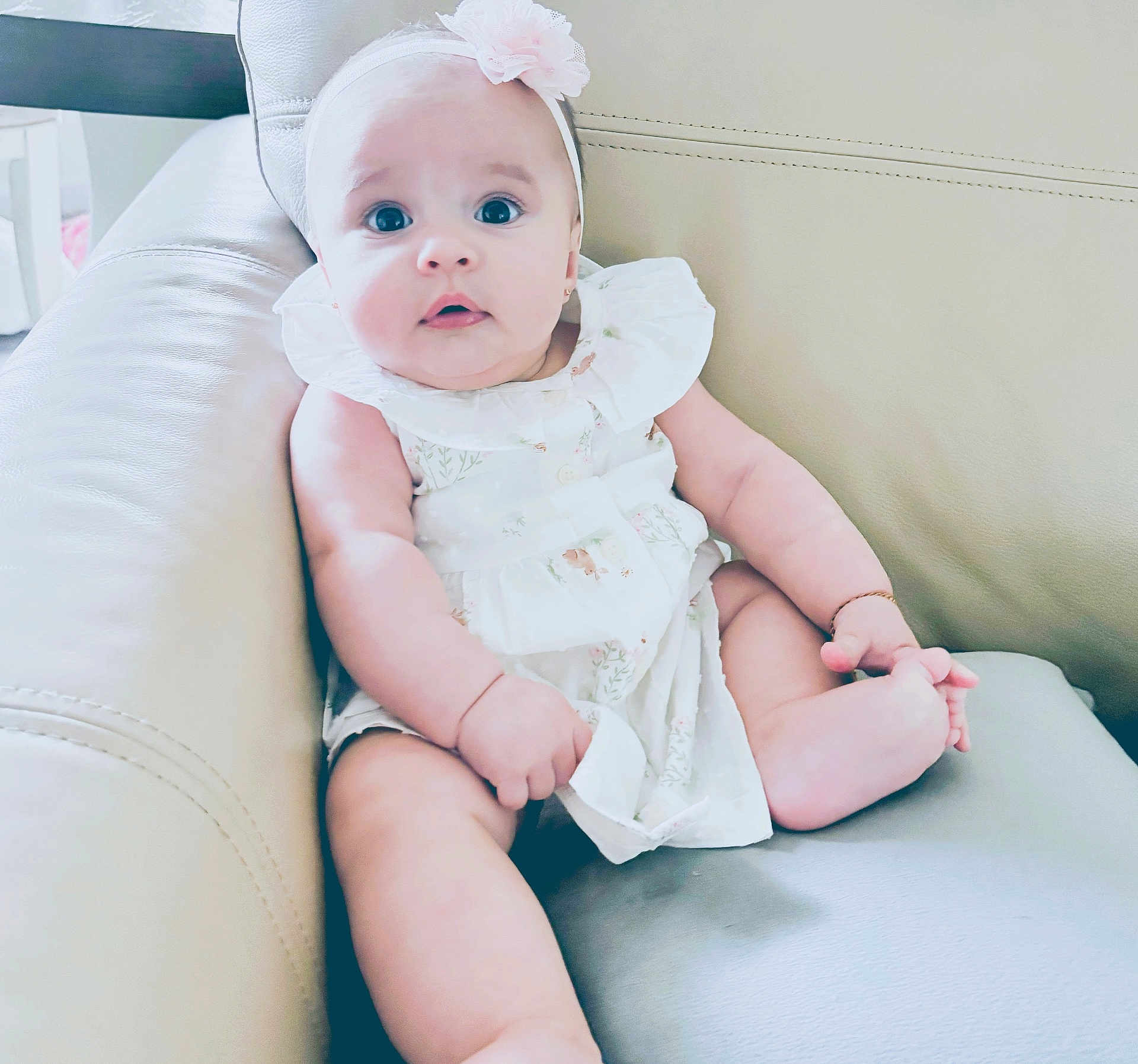 Valentina joined the competition — help win amazing prizes! baby, child, infant, cute, sitting, white_dress, headband, flower, leather_couch, indoor, curious, chubby, skin, foot, hand, portrait, person, young, furniture, comfortable