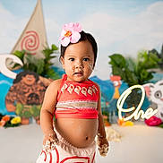 Brianna is registered to the contest to win money with this photo: toddler, child, birthday, cake, flower, headband, red_clothing, smiling, indoor, celebration, moana_theme, decorations, colorful, cartoon_characters, background, plants, happy, portrait, party, one_year_old