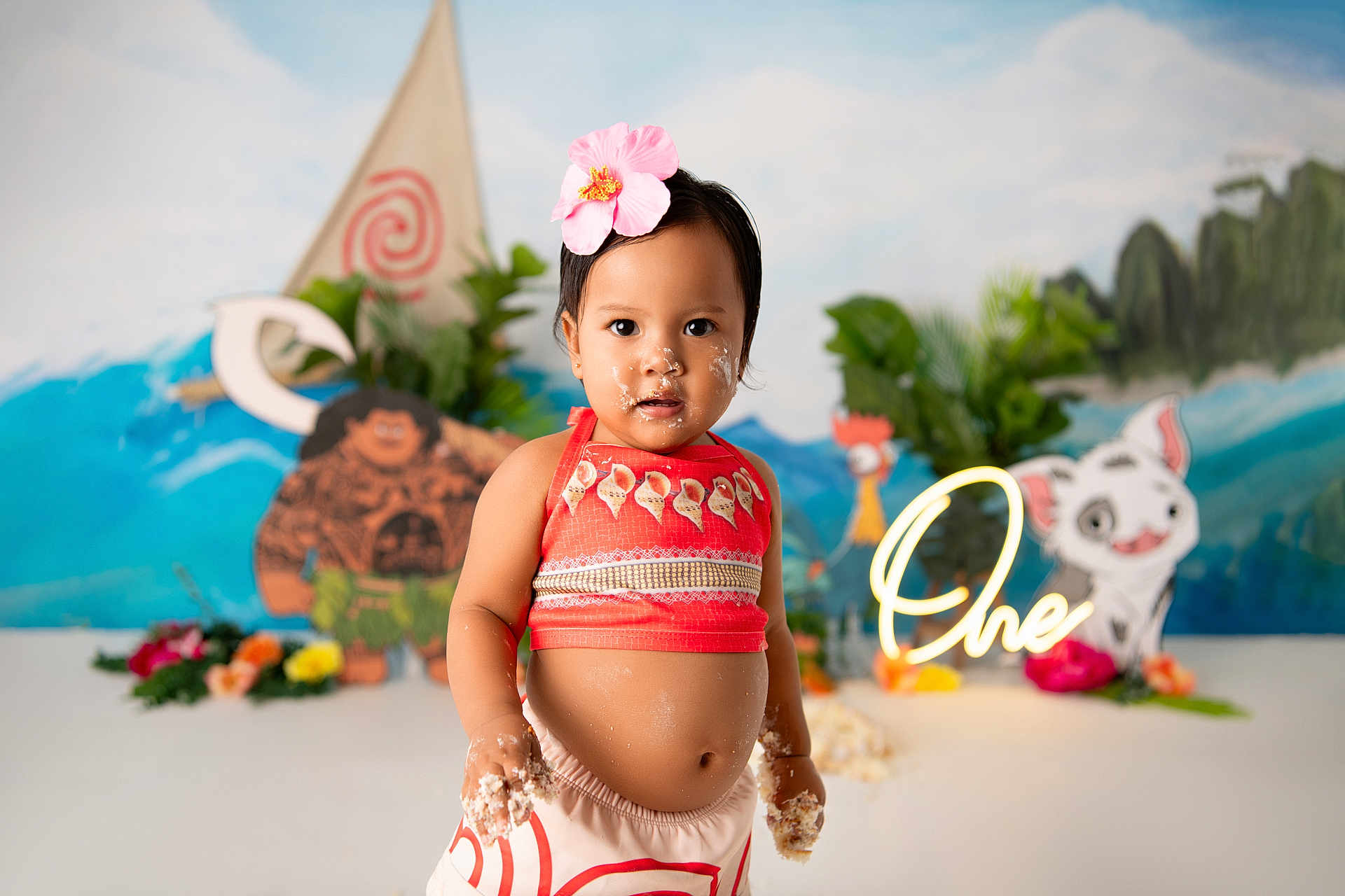 Brianna is registered to the contest to win money with this photo: toddler, child, birthday, cake, flower, headband, red_clothing, smiling, indoor, celebration, moana_theme, decorations, colorful, cartoon_characters, background, plants, happy, portrait, party, one_year_old