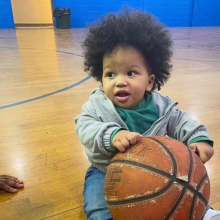 Nazir Anderson is registered to the contest to win money with this photo: ball, basketball, casual_clothing, child, curly_hair, expression, floor, gym, hand, hoodie, indoor, indoor_sports, jeans, person, playful, shoe, sports, toddler, wooden_floor, young_child