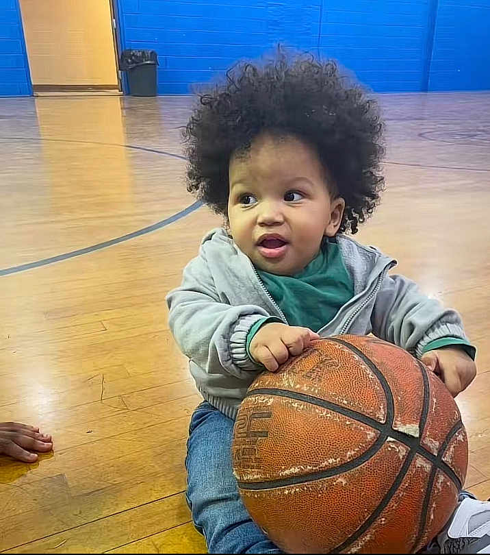 Nazir Anderson is registered to the contest to win money with this photo: toddler, child, basketball, indoor, gym, floor, curly_hair, casual_clothing, hoodie, jeans, shoe, hand, sports, playful, young_child, person, wooden_floor, ball, expression, indoor_sports