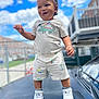 toddler, child, smiling, playful, outdoor, clouds, blue_sky, car, sneakers, shorts, tshirt, urban, fence, daylight, standing, person, happy, young_child, fashion, portrait
