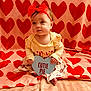 baby, blanket, bow, child, cozy, cute, cutie_pie_sign, headband, heart_pattern, heart_shaped_sign, infant, onesie, pajamas, pink_blanket, portrait, red_hearts, sign_with_text, sitting, small_feet, soft_texture