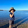Elias joined the competition — help win amazing prizes! child, toddler, hat, water, lake, sky, blue_sky, outdoor, nature, summer, shoes, shorts, tshirt, standing, reflection, cityscape, pebble_beach, sunlight, shadow, portrait