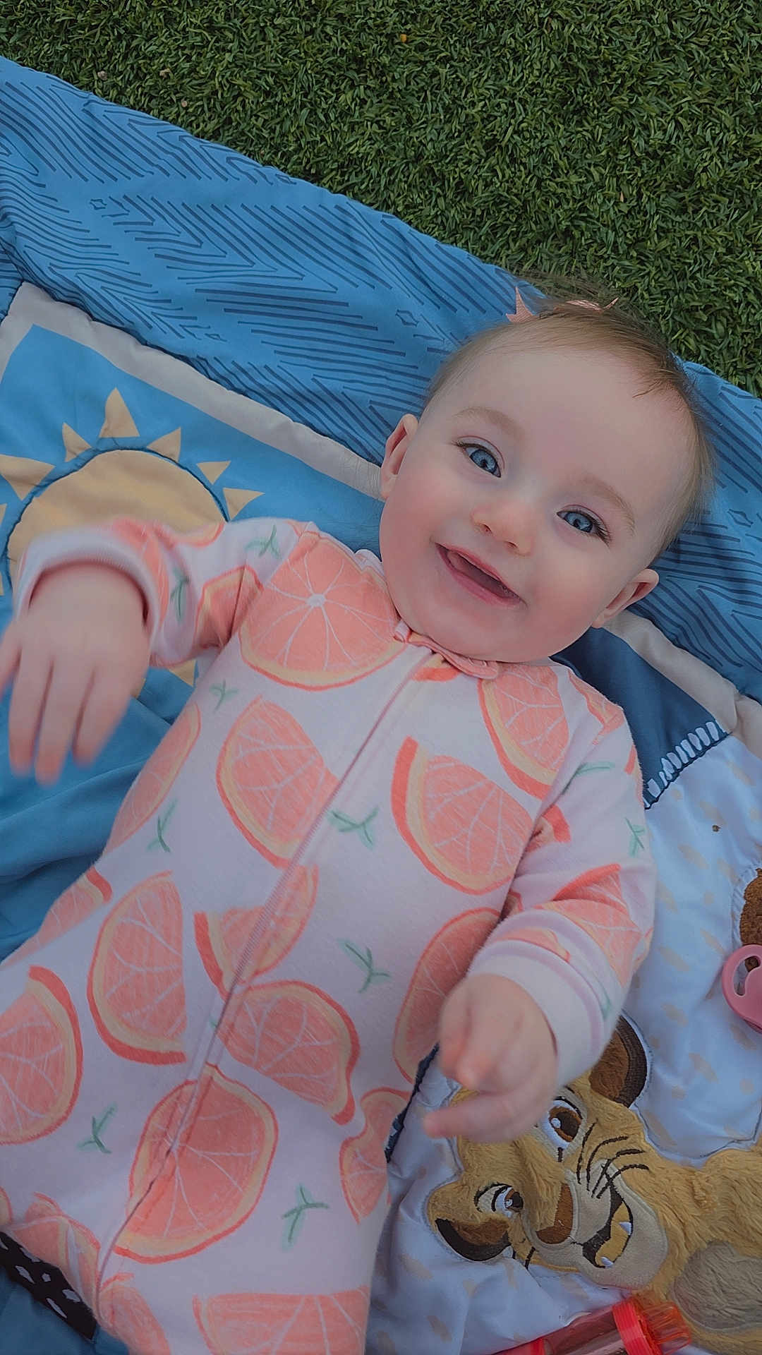 Lainey is registered to the contest to win money with this photo: baby, infant, smile, happy, onesie, orange_slice_pattern, blanket, grass, blue, playmat, portrait, face, cheek, eyes, hand, toy, lion_plush, outdoor, cute, laying_down