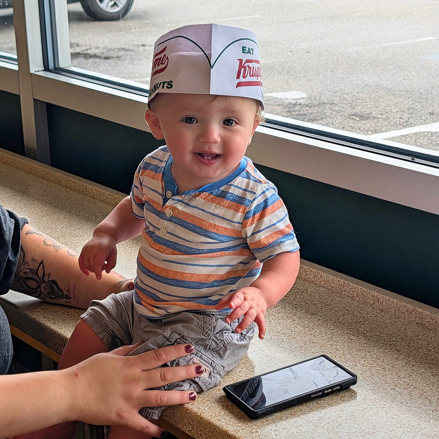 Leo joined the competition — help win amazing prizes! baby, child, toddler, smiling, sitting, portrait, indoor, restaurant, diner, counter, window, parking_lot, paper_hat, striped_shirt, shorts, barefoot, adult, hand, smartphone, vehicle