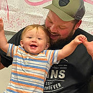 Leo is registered to the contest to win money with this photo: baby, child, infant, father, parent, family, smiling, happy, laughing, portrait, indoor, celebration, striped_shirt, baseball_cap, beard, arms_raised, sitting, fabric_backdrop, hearts, family_photo