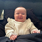 Layla joined the competition — help win amazing prizes! baby, smiling, white_sweater, frills, blanket, cushion, phone, indoor, child, happy, portrait, face, hands, cozy, infant, cute, warm, resting, soft, clothing