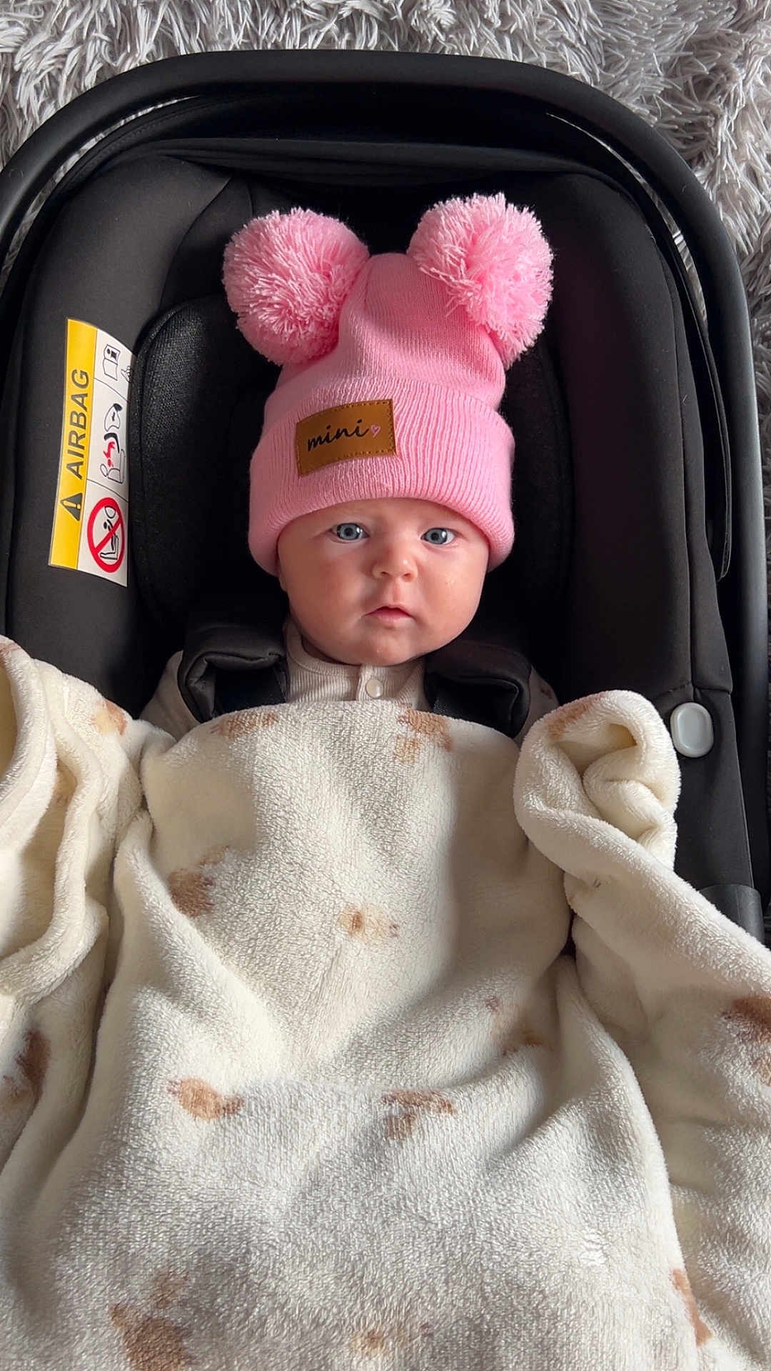 Layla is registered to the contest to win money with this photo: baby, infant, car_seat, blanket, pink_hat, pom_poms, face, blue_eyes, cozy, warm, indoor, soft_texture, cute, child, person, headwear, smiling, closeup, car_seat_strap, floor_rug