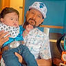 Jr is registered to the contest to win money with this photo: baby, beard, belt, cap, child, clothing, family, father, holding, home, indoor, jeans, parent, portrait, pouting, shirt, sitting, smiling, table, television