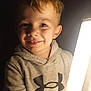 child, smiling, hoodie, blond_hair, under_armour, indoor, portrait, light_source, happy, casual_clothing, young, cute, face, eyes, person, warm_lighting, dark_background, close_up, childhood, expression