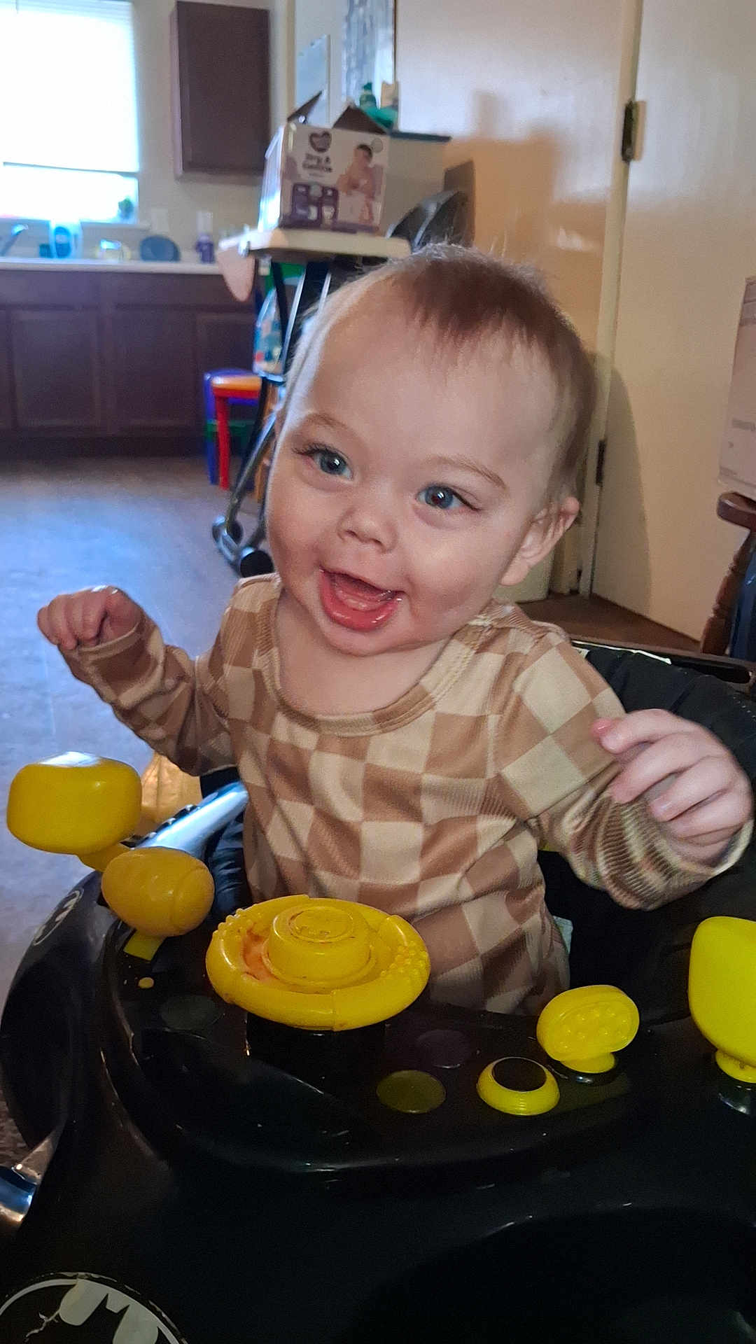 Chase is registered to the contest to win money with this photo: toddler, child, baby, walker, toy, yellow, black, checkered_shirt, smiling, happy, indoor, kitchen, furniture, floor, face, person, cute, playful, young_child, home