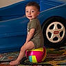 child, toddler, boy, sitting, smiling, portrait, toy_car, toy_vehicle, toy_wheel, colorful_ball, indoor, playroom, carpet, blue, green_shirt, barefoot, playtime, vehicle, wheel, happy