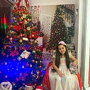 Nathaly Roque joined the competition — help win amazing prizes! child, girl, christmas_tree, holiday_decorations, santa_claus, gift_box, dress, flower_headband, indoor, festive, smiling, long_hair, floor, window, lights, ornaments, holiday, celebration, seasonal, decor