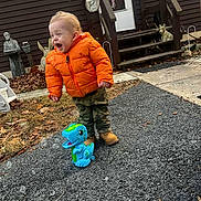 Kaleb is registered to the contest to win money with this photo: toddler, child, orange_jacket, laughing, toy, dinosaur, bubble_machine, outdoor, pavement, leaves, house, stairs, boots, camouflage_pants, statue, grass, play, happy, person, daylight