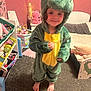 child, dinosaur_costume, barefoot, smile, indoor, carpet, toy_shelf, bedroom, childrens_room, plush_toy, pink_wall, furniture, playroom, young_child, cute, costume, person, feet, curly_hair, happy
