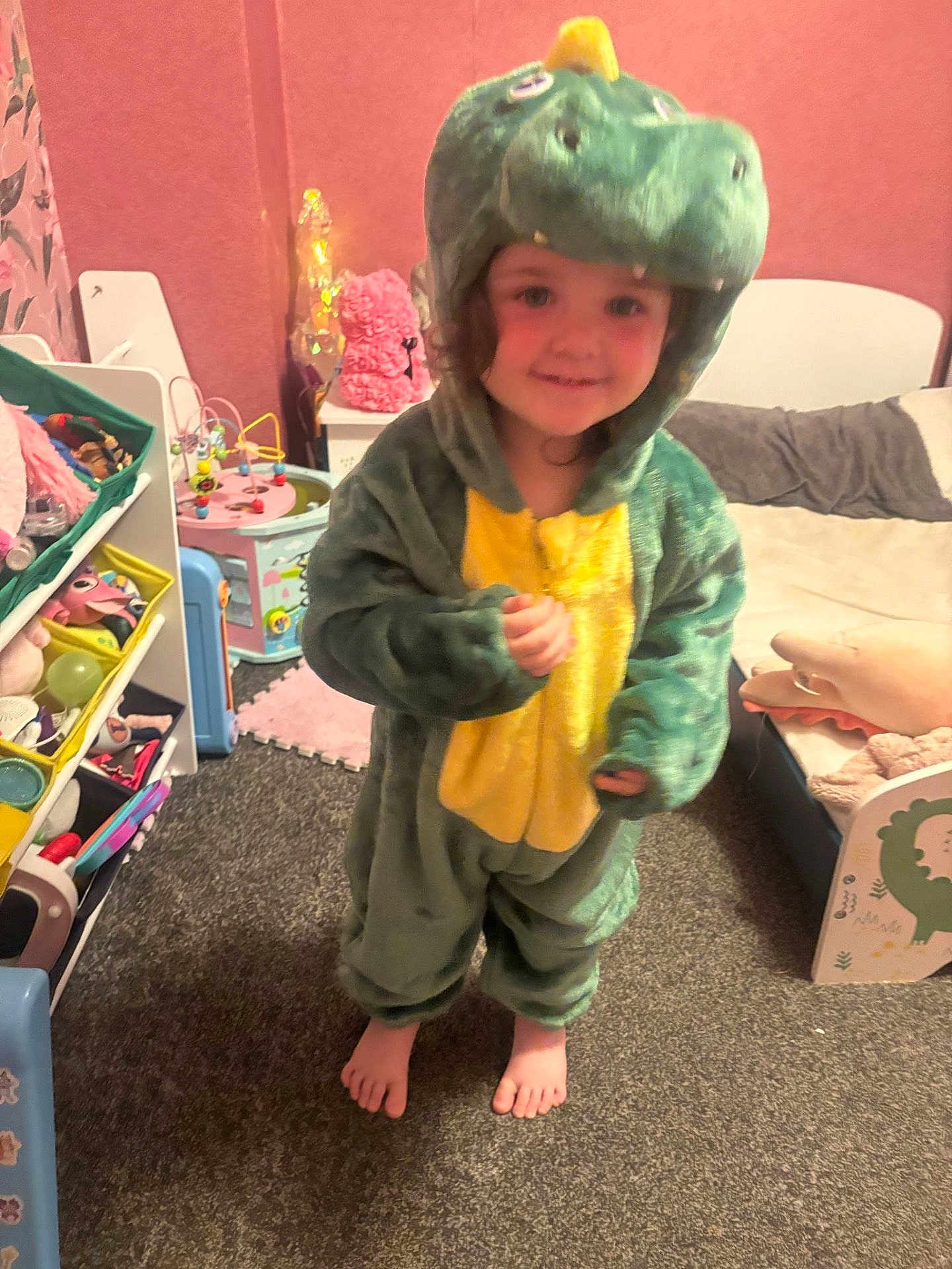 Danni joined the competition — help win amazing prizes! child, dinosaur_costume, barefoot, smile, indoor, carpet, toy_shelf, bedroom, childrens_room, plush_toy, pink_wall, furniture, playroom, young_child, cute, costume, person, feet, curly_hair, happy