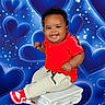 baby, child, smiling, red_shirt, sneakers, pedestal, portrait, studio_photography, blue_background, hearts, stars, happy, cute, curly_hair, clothing, sitting, infant, person, fashion, joyful