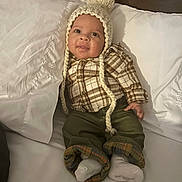 William is registered to the contest to win money with this photo: baby, child, infant, hat, knitted_hat, pom_pom, plaid_shirt, green_pants, socks, bed, pillow, white_bedding, smiling, cute, cozy, indoor, portrait, person, clothing, happy