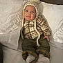 baby, child, infant, hat, knitted_hat, pom_pom, plaid_shirt, green_pants, socks, bed, pillow, white_bedding, smiling, cute, cozy, indoor, portrait, person, clothing, happy