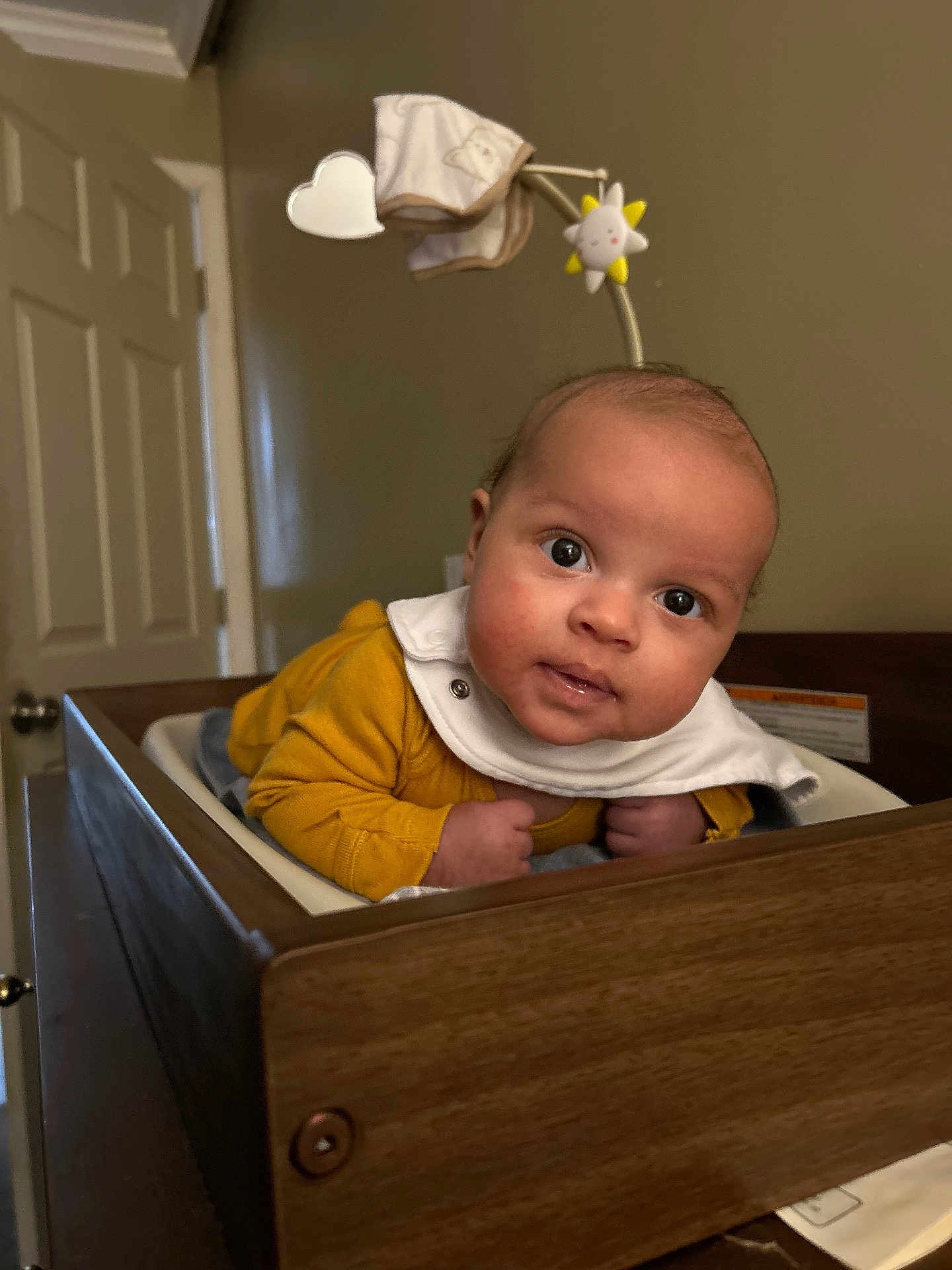 William is registered to the contest to win money with this photo: baby, infant, wooden_crib, mobile_toy, yellow_clothing, curious_expression, indoors, face, head, person, child, furniture, room, door, wall, light, white_bib, close_up, portrait, cute