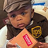 Dream joined the competition — help win amazing prizes! baby, child, costume, ups_uniform, package, fragile_label, hat, delivery, portrait, indoor, person, cute, curious, hands, seated, small_box, brown_clothing, earrings, expression, closeup