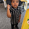 child, toddler, boy, striped_shirt, black_shoes, carpet, indoor, table, chair, tablecloth, person, standing, serious, gesture, finger_raised, curly_hair, young, cute, expression, floor
