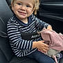 child, toddler, car_seat, striped_shirt, blonde_hair, blue_eyes, smiling, toy, blanket, seatbelt, car_interior, footwear, casual_clothing, happy, portrait, indoor, cute, young_child, person, sitting
