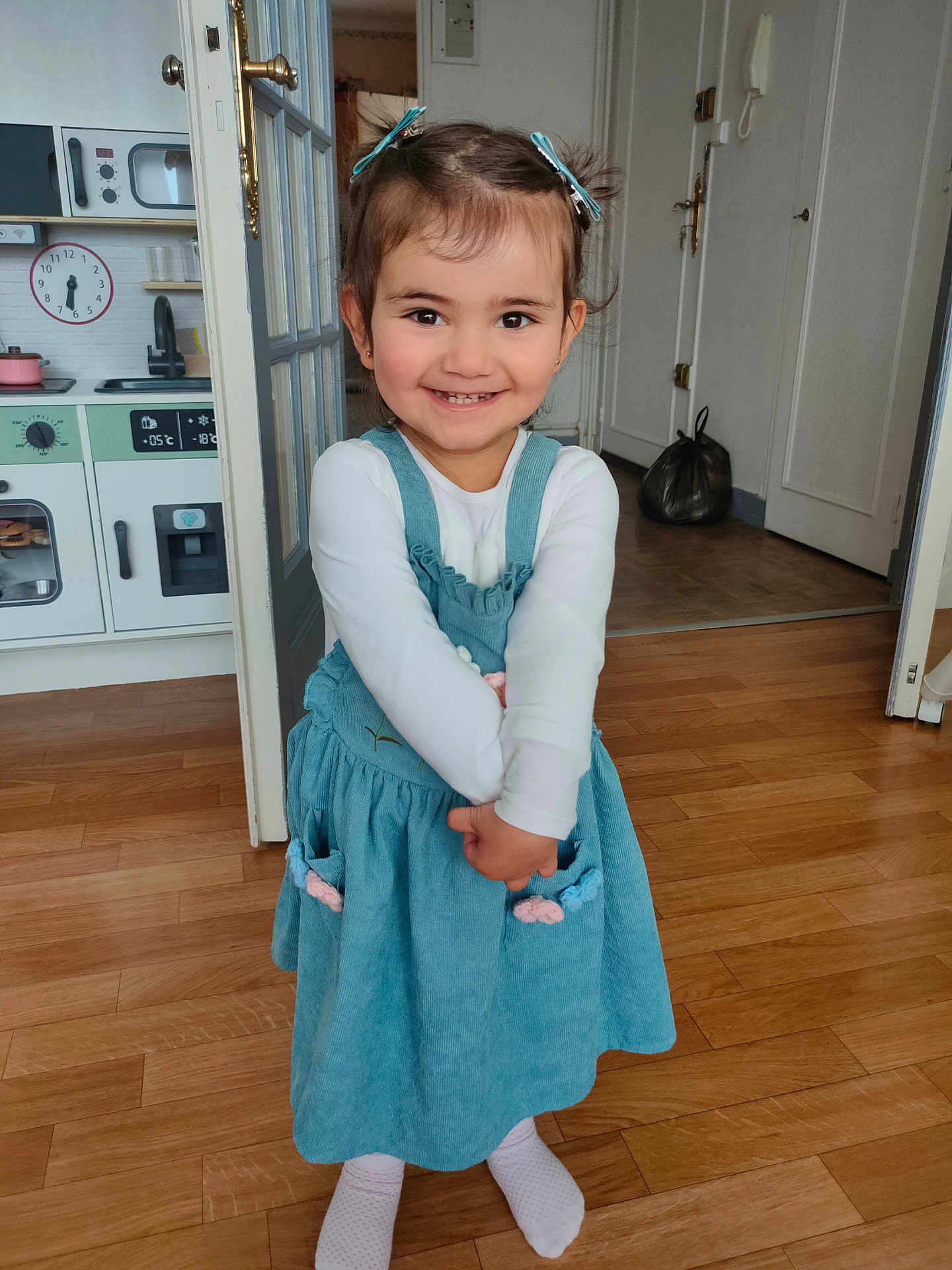 Melissa participe au concours pour gagner de l'argent avec cette photo : child, girl, smiling, indoor, wooden_floor, blue_dress, hair_clips, long_sleeve_shirt, play_kitchen, doorway, home_interior, happy, person, feet, socks, flooring, room, toy_kitchen, young_child, casual_clothing