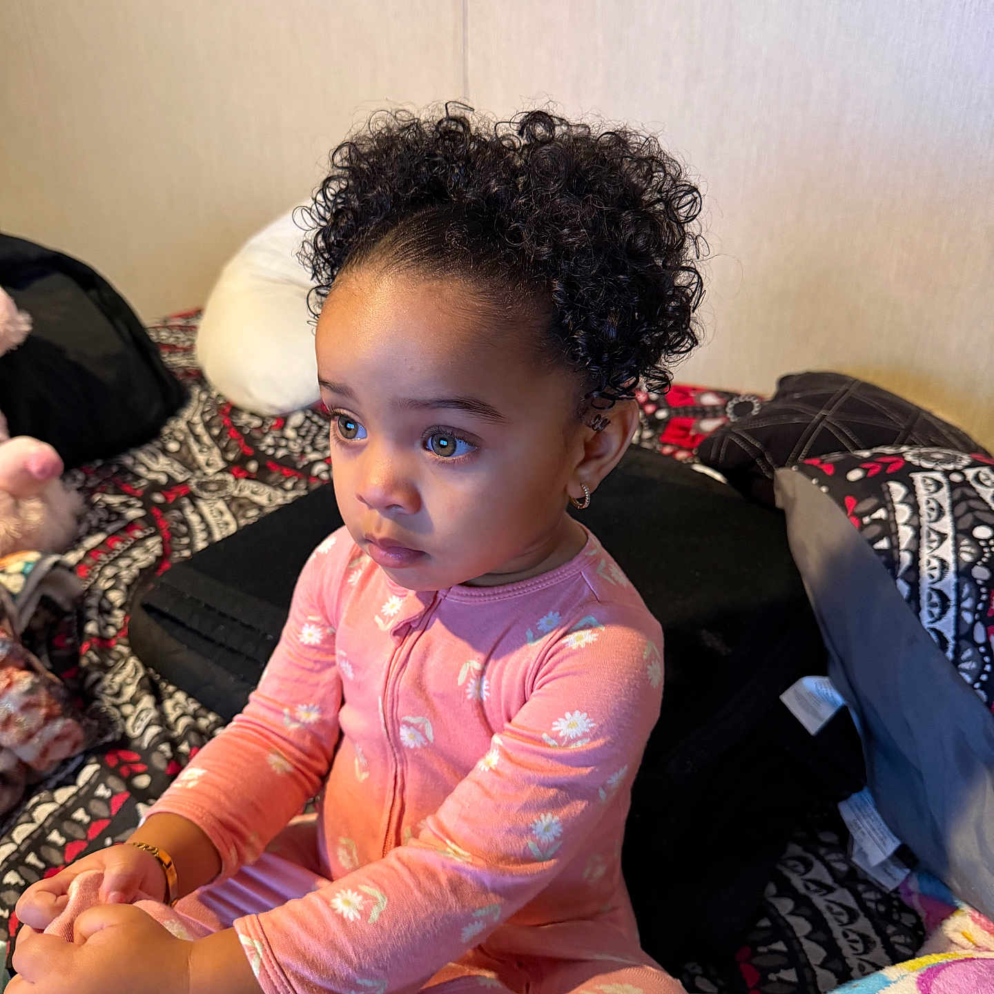 Jahira is registered to the contest to win money with this photo: toddler, child, curly_hair, pink_onesie, bed, blanket, indoor, patterned_bedspread, earrings, expression, cute, sitting, young_child, face, person, portrait, cozy, clothing, home, soft_lighting