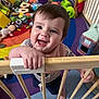 toddler, child, smiling, playpen, wooden_bars, toys, colorful, indoor, baby, happy, face, hand, play_area, floor, clothing, bib, expression, cute, person, home