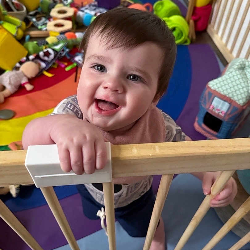 John joined the competition — help win amazing prizes! baby, bib, child, clothing, colorful, cute, expression, face, floor, hand, happy, home, indoor, person, play_area, playpen, smiling, toddler, toys, wooden_bars