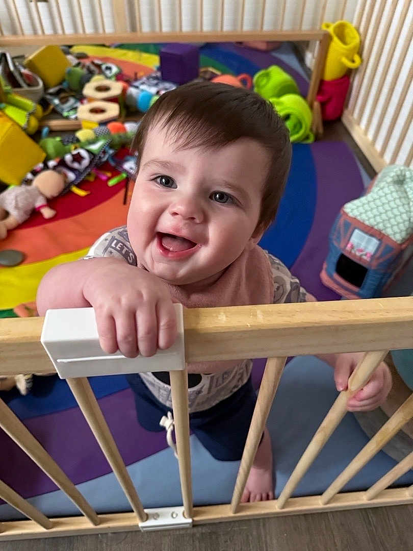 John joined the competition — help win amazing prizes! toddler, child, smiling, playpen, wooden_bars, toys, colorful, indoor, baby, happy, face, hand, play_area, floor, clothing, bib, expression, cute, person, home