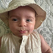 Theodore is registered to the contest to win money with this photo: baby, child, hat, bucket_hat, striped_clothing, beige, close_up, portrait, cute, infant, pout, face, blue_eyes, sleeveless, buttoned_shirt, quilt, patterned_background, skin, person, lying_down