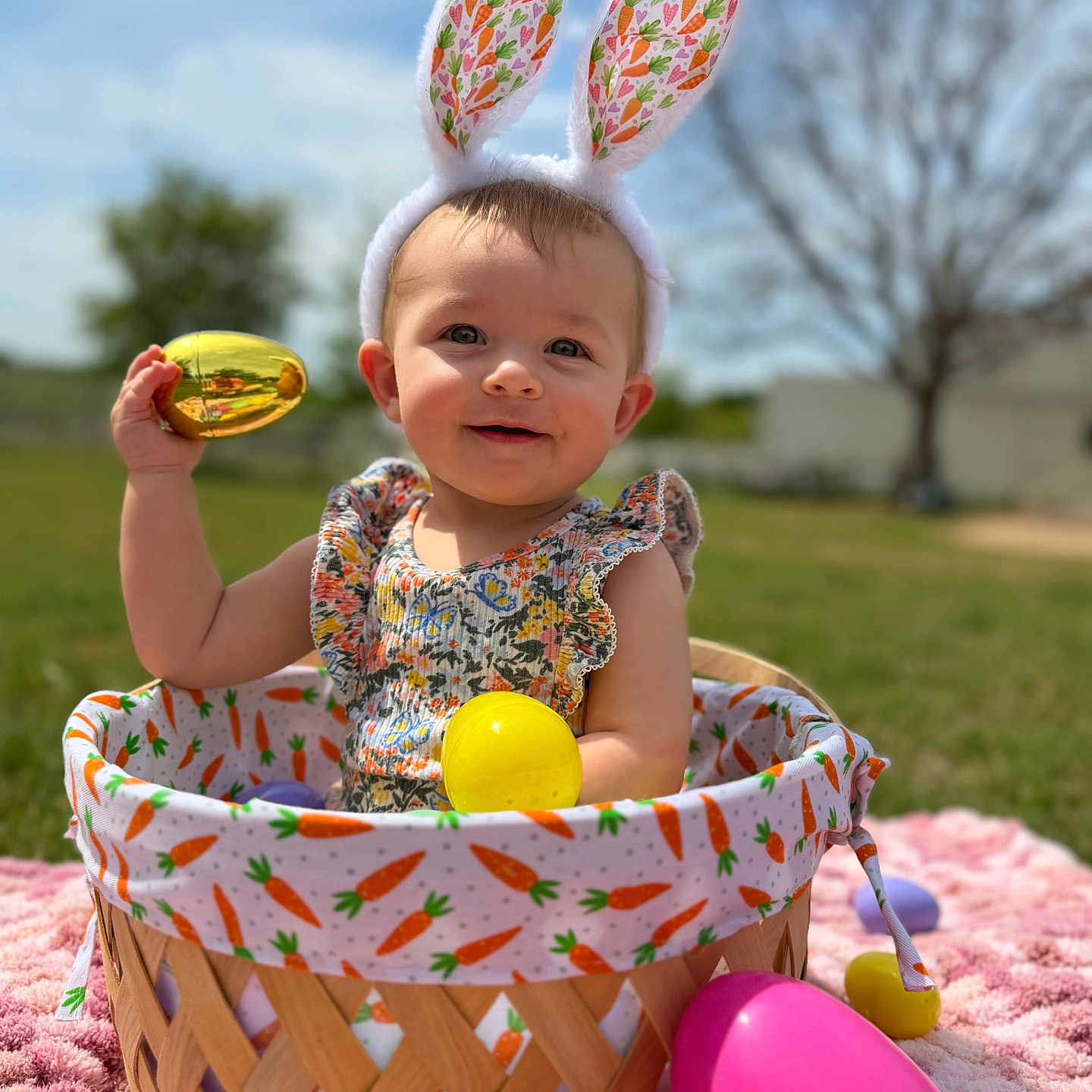 Helena is registered to the contest to win money with this photo: baby, basket, blue_sky, bunny_ears, child, colorful, cute, easter_eggs, fabric, grass, headwear, holding, outdoor, patterned, pink_blanket, playful, smiling, spring, sunny, tree